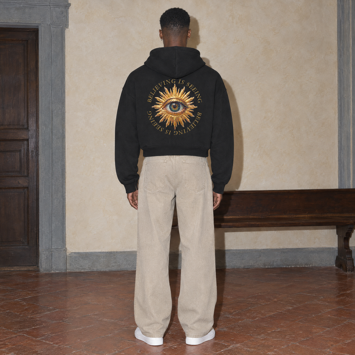 Man seen from behind wearing the black Believing full zip hoodie by Serenos, golden eye back print reading believing is seeing, cropped boxy fit