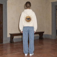 Woman seen from behind wearing the cream Believing full zip hoodie by Serenos, golden eye back print reading believing is seeing, cropped boxy fit