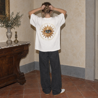 Woman seen from behind wearing the white Believing oversized t-shirt by Serenos, golden eye back print reading believing is seeing