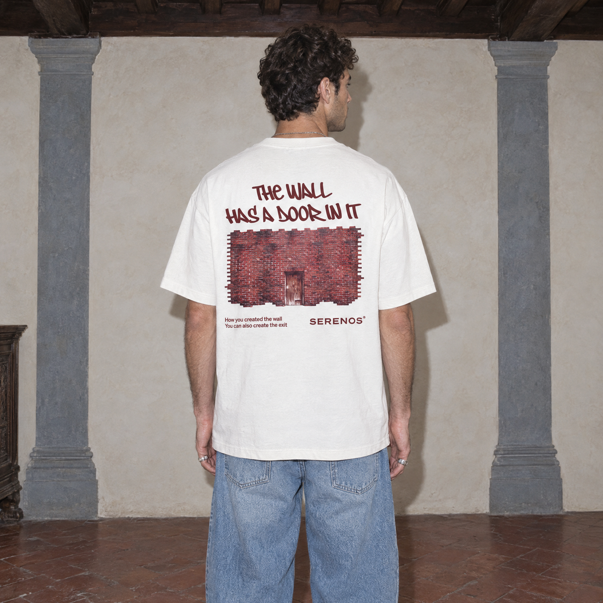 Man wearing the white Door t-shirt from behind, burgundy back print reading The wall has a door in it above a brick wall photo