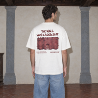 Man wearing the white Door t-shirt from behind, burgundy back print reading The wall has a door in it above a brick wall photo
