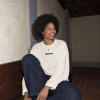 Lifestyle shot of a woman laughing on a walnut bench in a Florentine palazzo wearing the cream SERENOS Improvising long sleeve tee