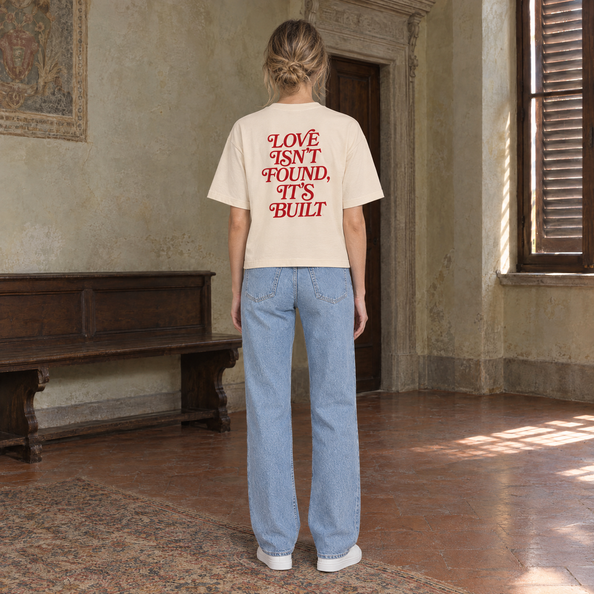 Woman seen from behind wearing the SERENOS Love T-Shirt in apricot, back print in red serif reading Love isn't found, it's built