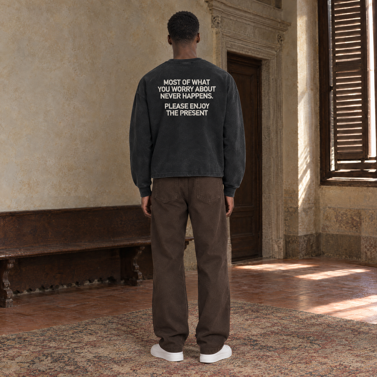 Man seen from behind wearing the SERENOS Never Happens long sleeve in washed black, back print reading Most of what you worry about never happens, please enjoy the present