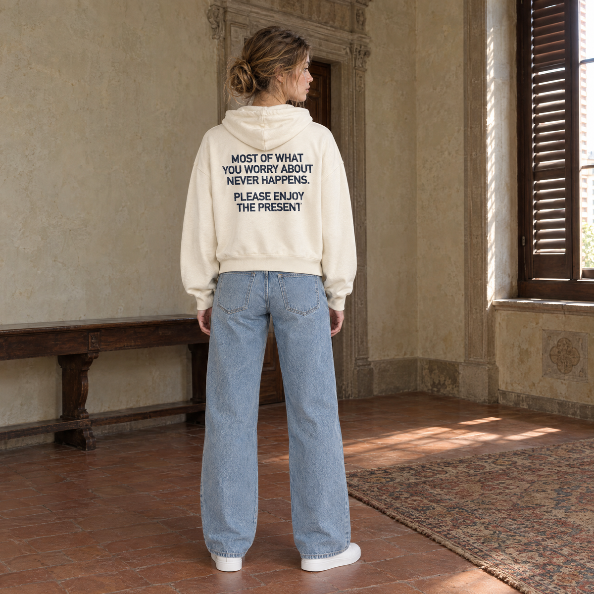 Woman seen from behind wearing the SERENOS Never Happens oversized boxy hoodie in apricot, back print reading Most of what you worry about never happens, please enjoy the present, cropped boxy fit