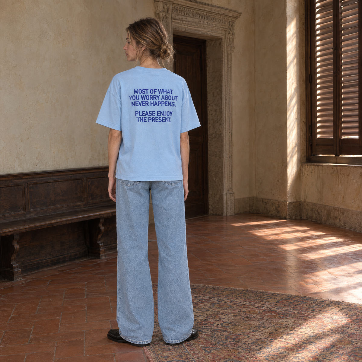 Woman wearing the SERENOS Never Happens T-Shirt in blue, back print reading Most of what you worry about never happens, please enjoy the present