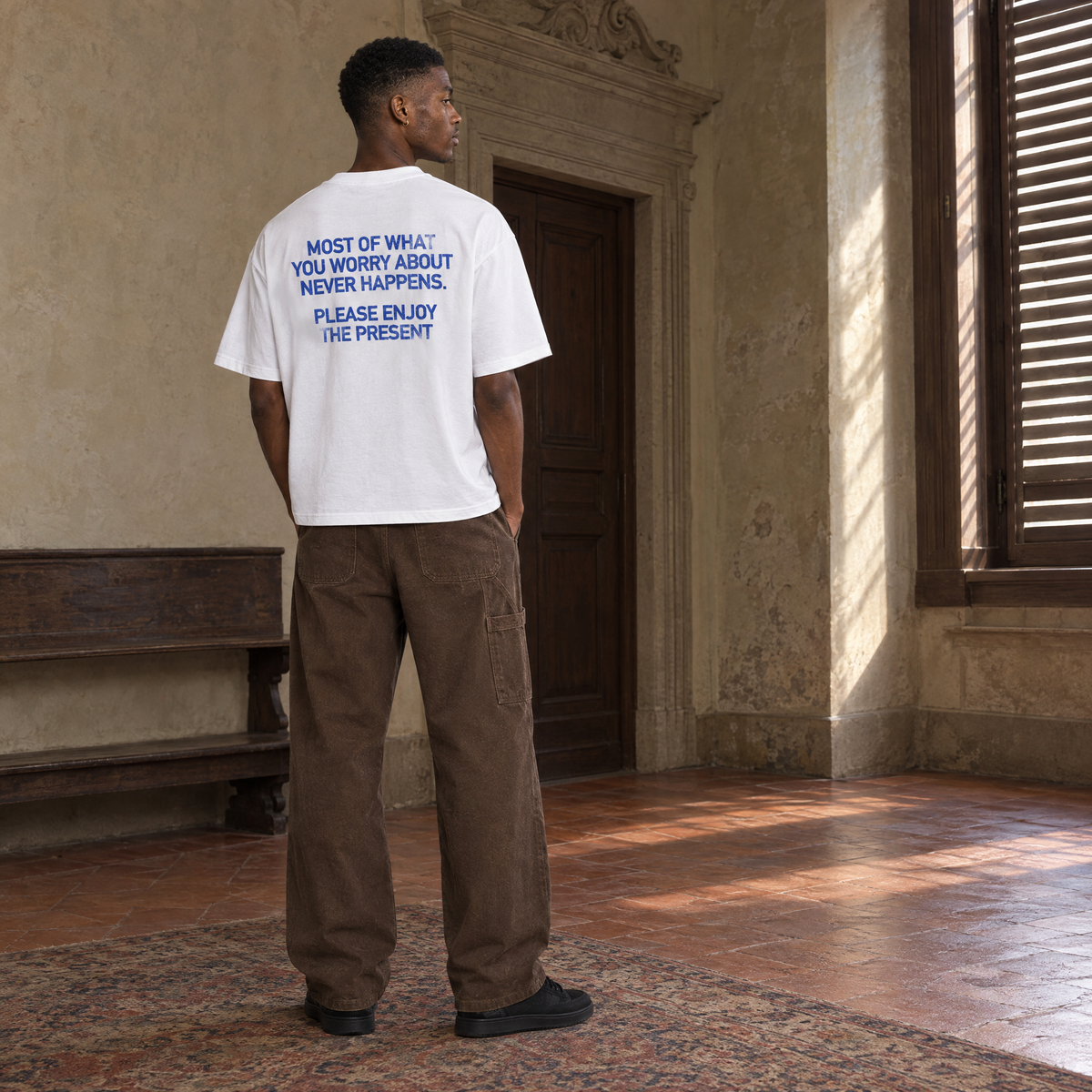 Man wearing the SERENOS Never Happens T-Shirt in white, back print reading Most of what you worry about never happens, please enjoy the present