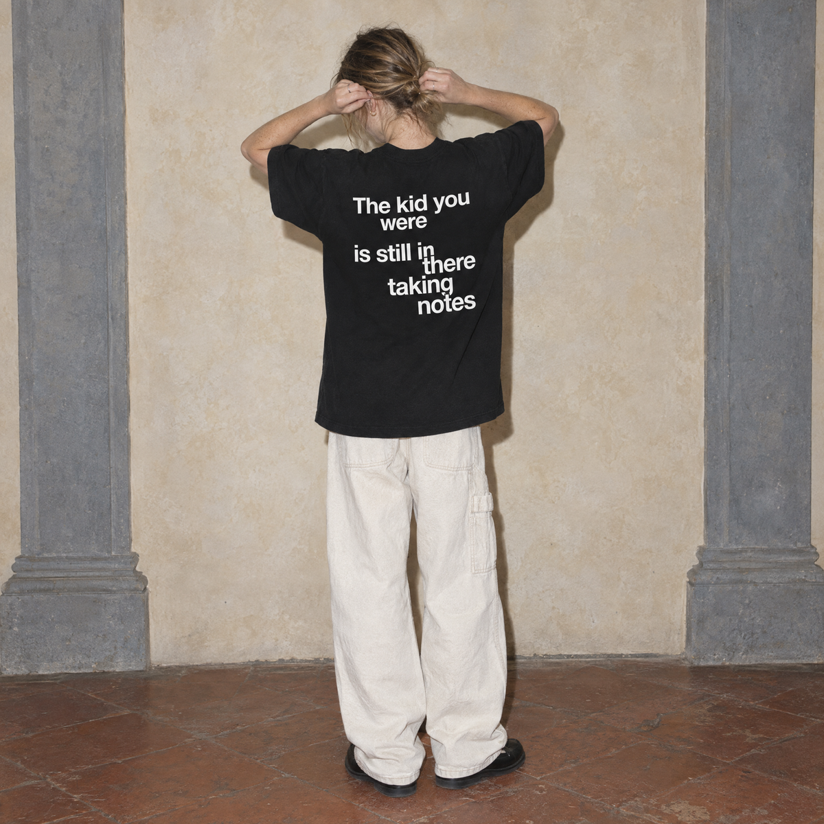 Woman seen from behind wearing the black Notes oversized t-shirt by Serenos, back print reading the kid you were is still in there taking notes