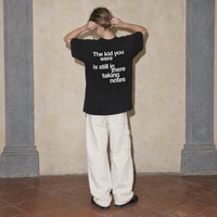 Woman seen from behind wearing the black Notes oversized t-shirt by Serenos, back print reading the kid you were is still in there taking notes