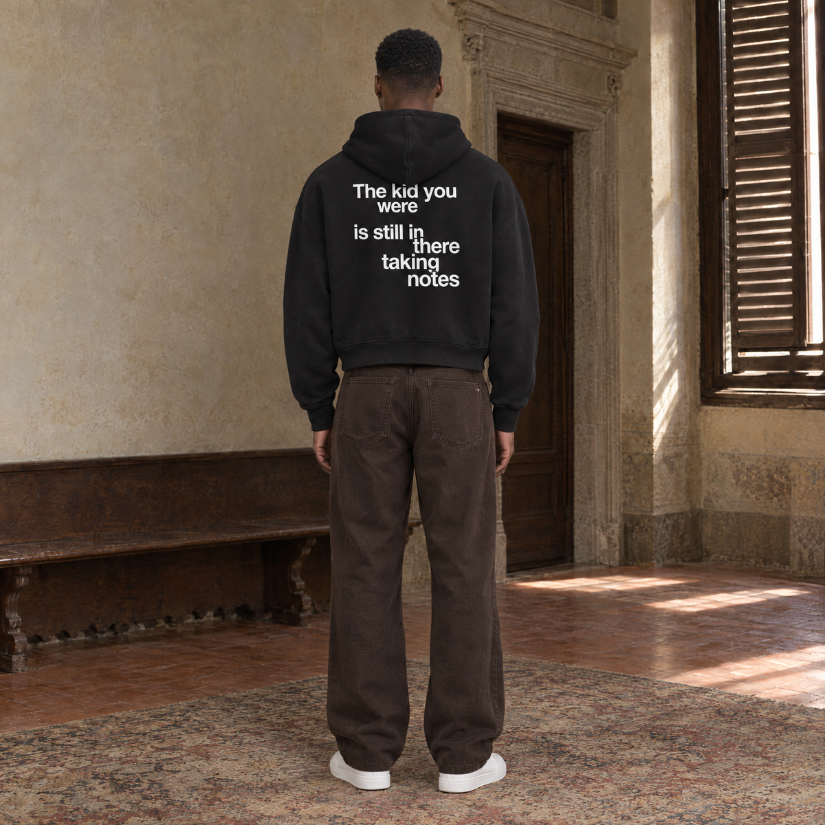 Man seen from behind wearing the SERENOS Notes zip hoodie in black, back print reading The kid you were is still in there taking notes, cropped boxy fit