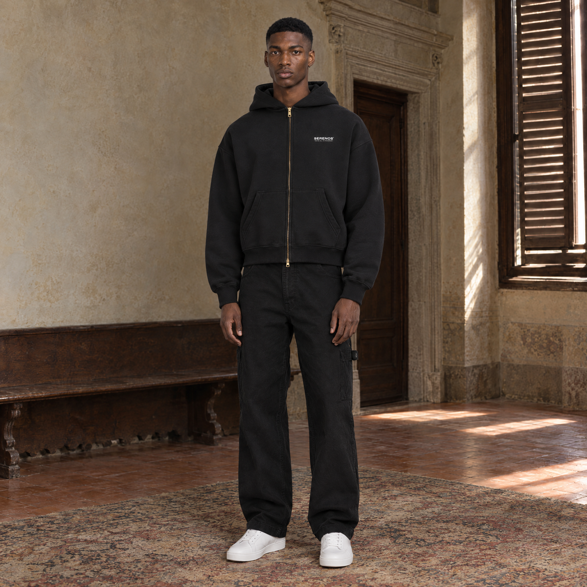 Man wearing the SERENOS Notes zip hoodie in black, front view zipped up with the brass zip and the small chest logo