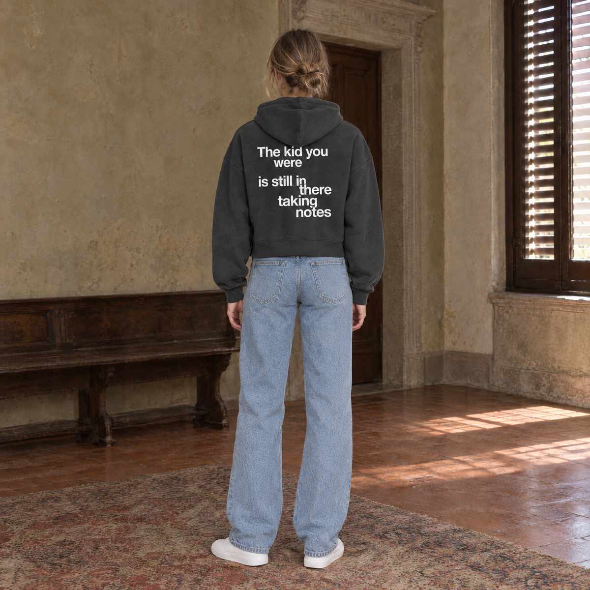 Woman seen from behind wearing the SERENOS Notes zip hoodie in dark gray, back print reading The kid you were is still in there taking notes, cropped boxy fit