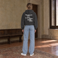 Woman seen from behind wearing the SERENOS Notes zip hoodie in dark gray, back print reading The kid you were is still in there taking notes, cropped boxy fit