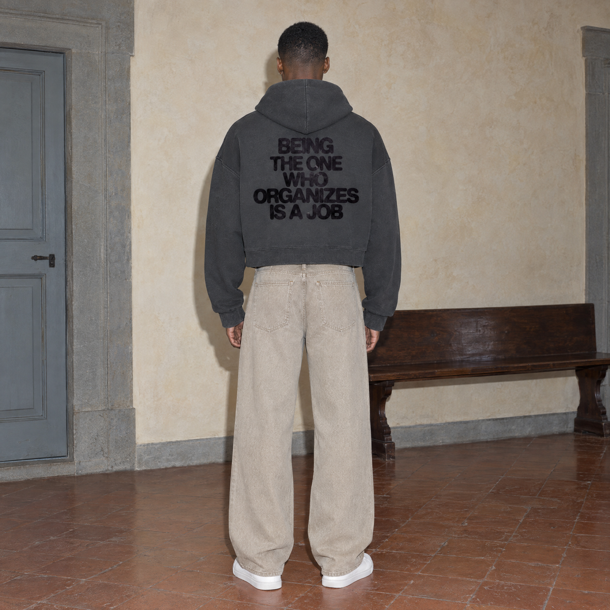 Man wearing the SERENOS Organizes oversized boxy hoodie in dark gray, seen from behind showing the Being The One Who Organizes Is A Job back print, cropped boxy fit