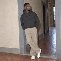 Man leaning on a pietra serena doorway wearing the SERENOS Organizes oversized boxy hoodie in dark gray in a Florentine palazzo room