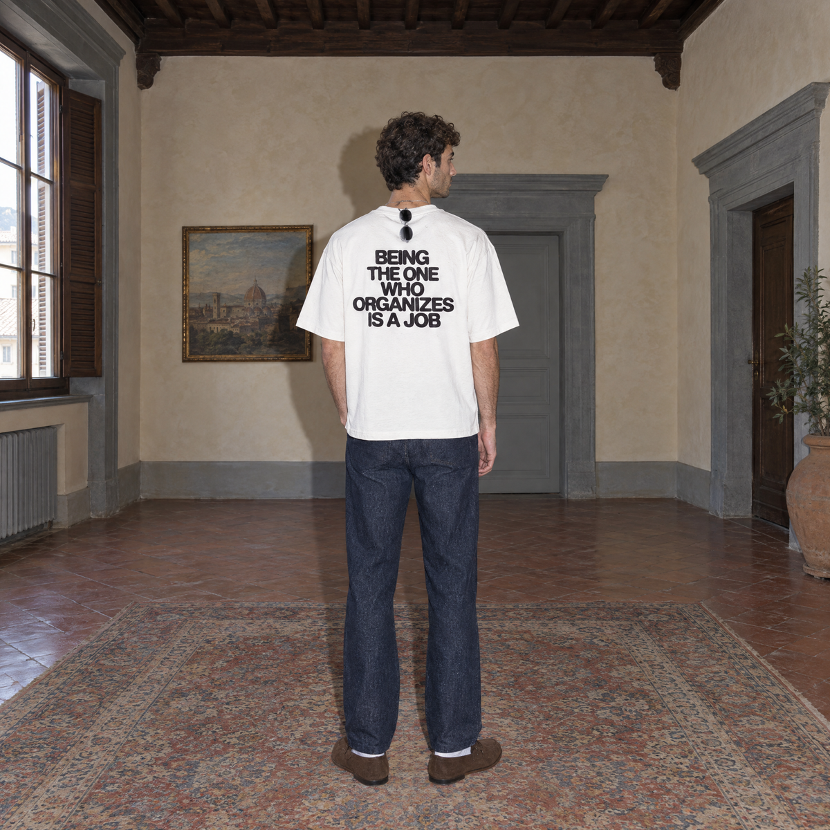 Man wearing the white Organizes t-shirt from behind, bold black back print reading Being the one who organizes is a job