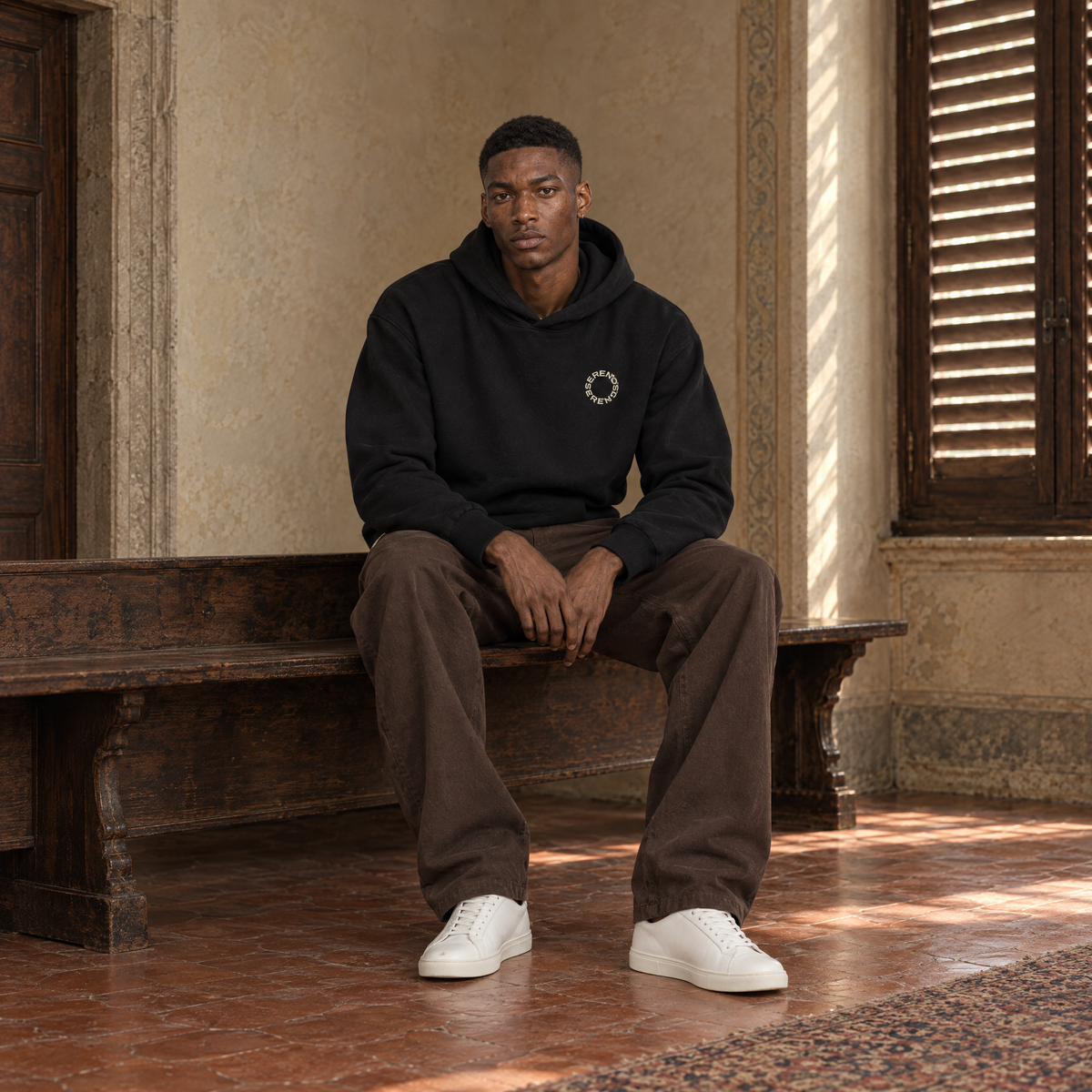 Man sitting on a bench in a Florentine palazzo wearing the SERENOS Shy oversized boxy hoodie in black