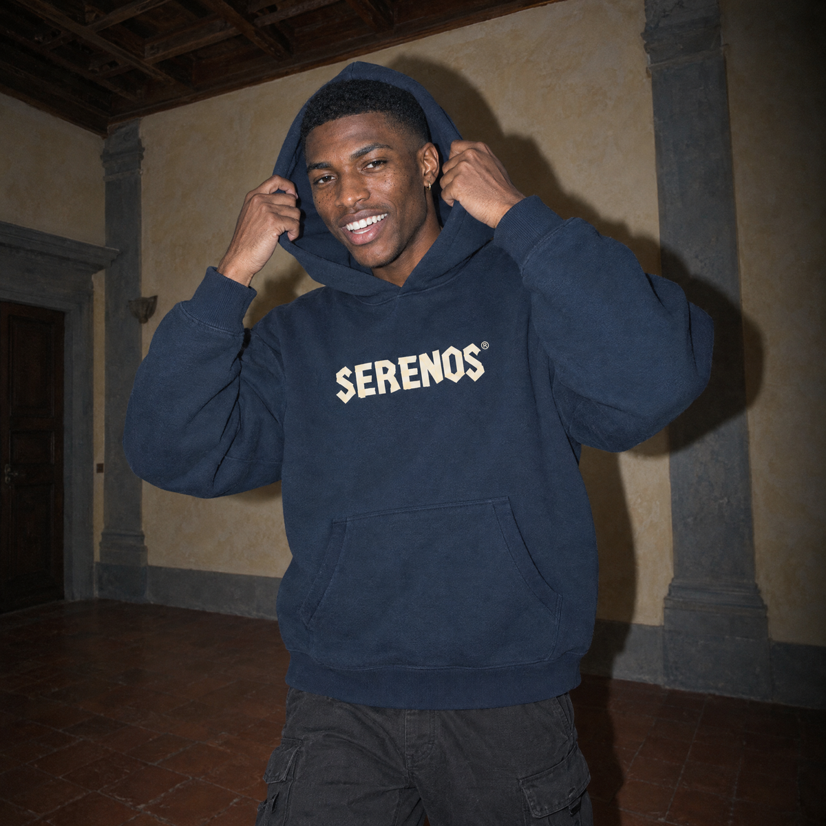 Lifestyle shot of a man pulling up the hood of the navy SERENOS Sun Above boxy hoodie in a Florentine palazzo