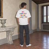Man wearing the white Therapist t-shirt from behind, multicoloured cut-paper back print reading My therapist knows about you