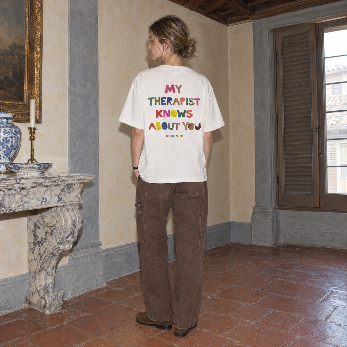 Woman wearing the white Therapist t-shirt from behind, multicoloured cut-paper back print reading My therapist knows about you