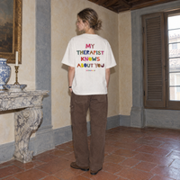 Woman wearing the white Therapist t-shirt from behind, multicoloured cut-paper back print reading My therapist knows about you
