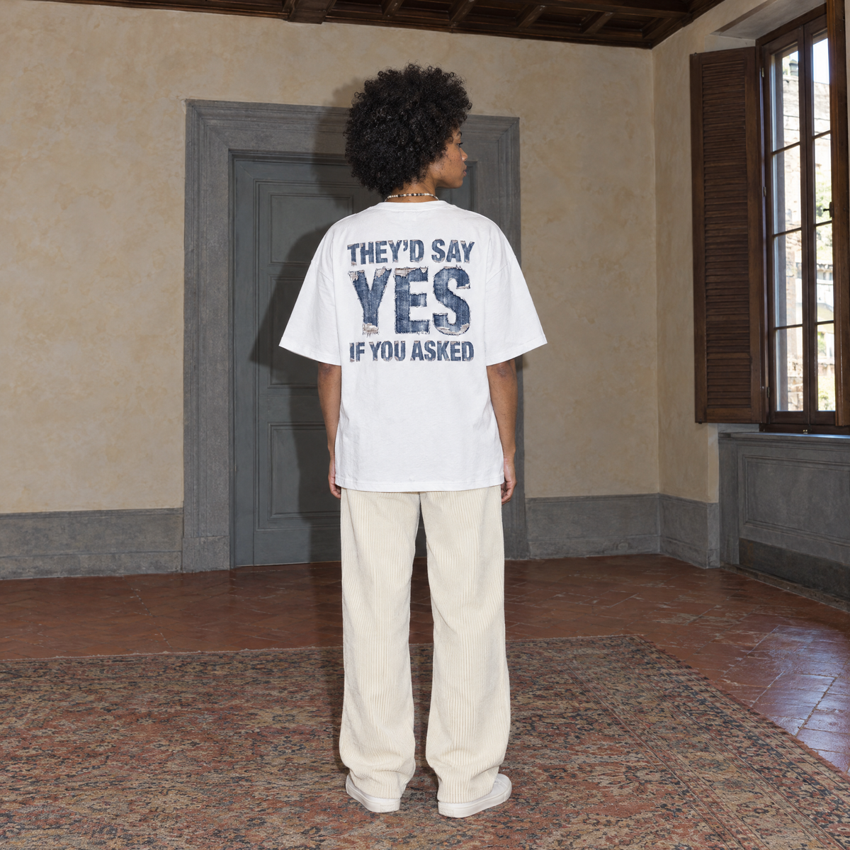 Woman wearing the white Yes t-shirt from behind, denim-textured back print reading They'd say yes if you asked